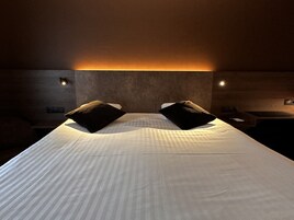 Habitación de hotel con una cama grande, dos almohadas negras, un cabecero de madera y luces fijadas en la pared.