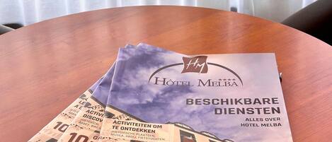 A brochure titled "Hotel Melba" on a wooden table with a window in the background.