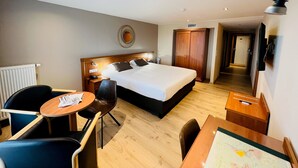 A hotel room with a bed, a desk, a chair, a round table, a TV, and a wardrobe. - Hotel Melba (Bastogne)