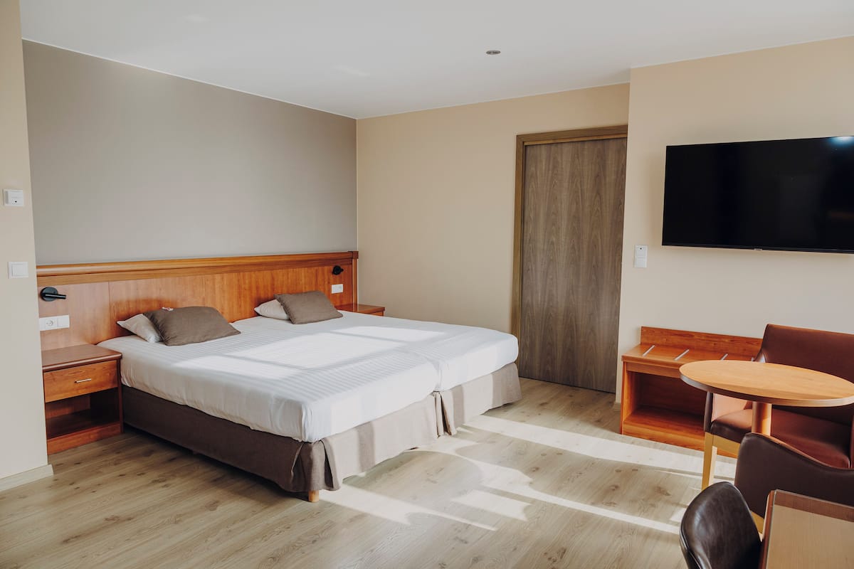 A hotel room with a double bed, a wooden nightstand, a desk with a chair, a television on the wall, and a wooden door.