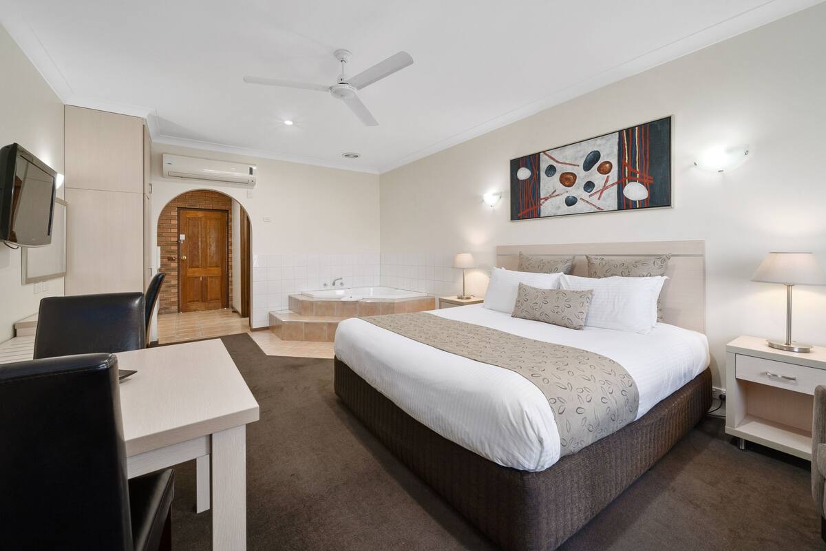 A hotel room with a bed, a desk, a chair, a TV, a ceiling fan, and a bathroom with a bathtub and shower.
