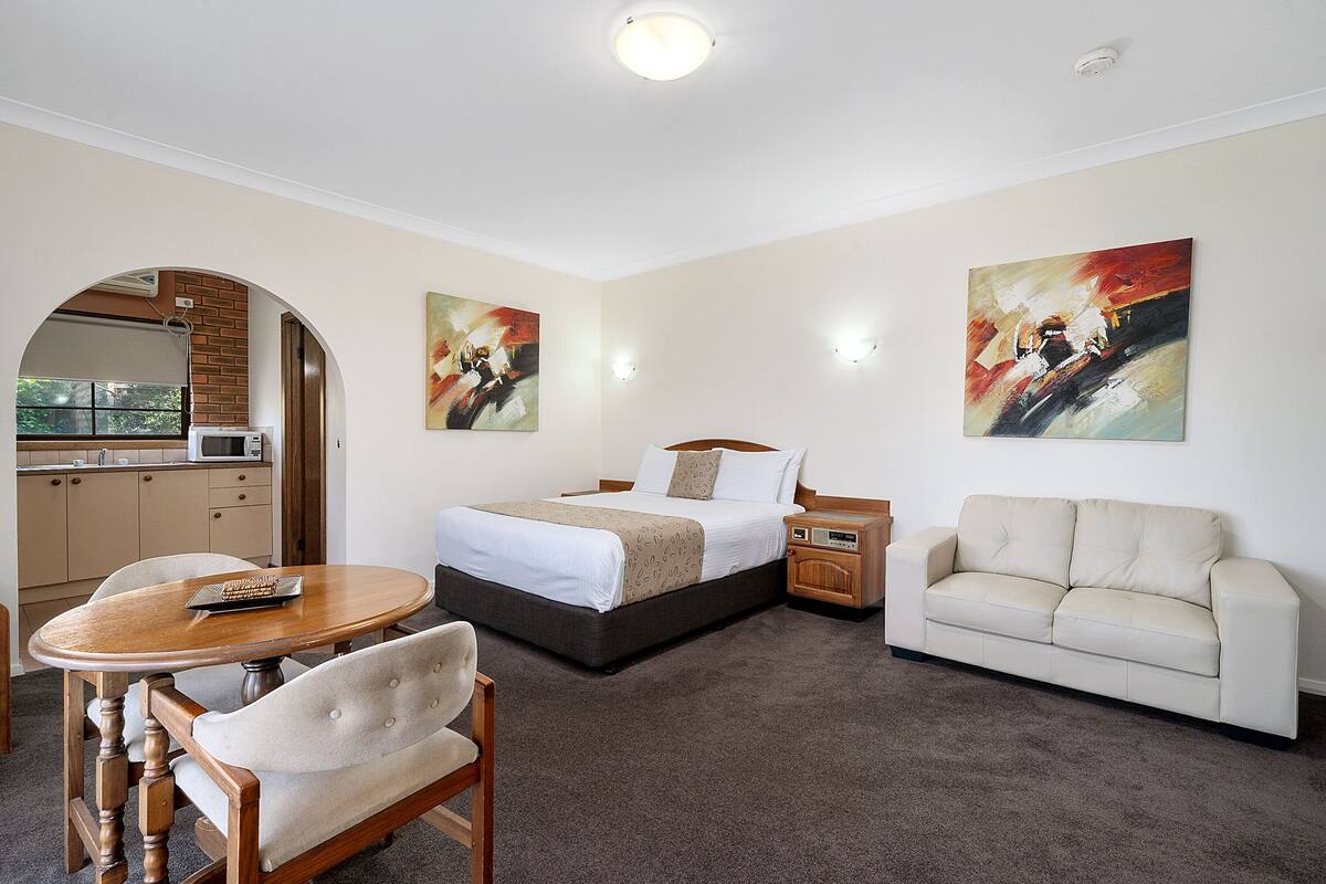 A hotel room with a bed, a sofa, a dining table, and a kitchen area visible through an archway.