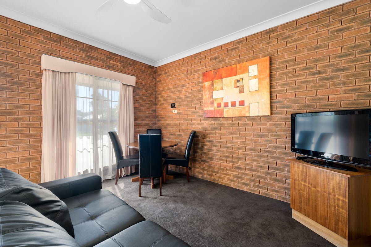 A living room with a brick wall, a flat-screen TV on a wooden stand, a sofa, a dining table with chairs, and a large window with curtains.