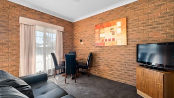 Una sala de estar con pared de ladrillo, un televisor de pantalla plana sobre un soporte de madera, un sofá, una mesa de comedor con sillas y un ventanal con cortinas.