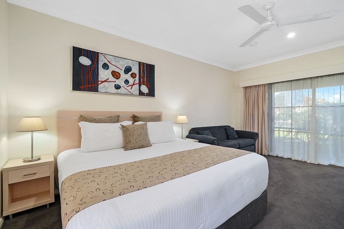 A hotel room with a bed, a sofa, a ceiling fan, a painting, and a window with curtains.