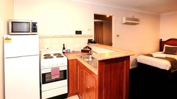 A compact kitchen with a microwave, stove, and sink, adjacent to a small bedroom with a bed and a nightstand.