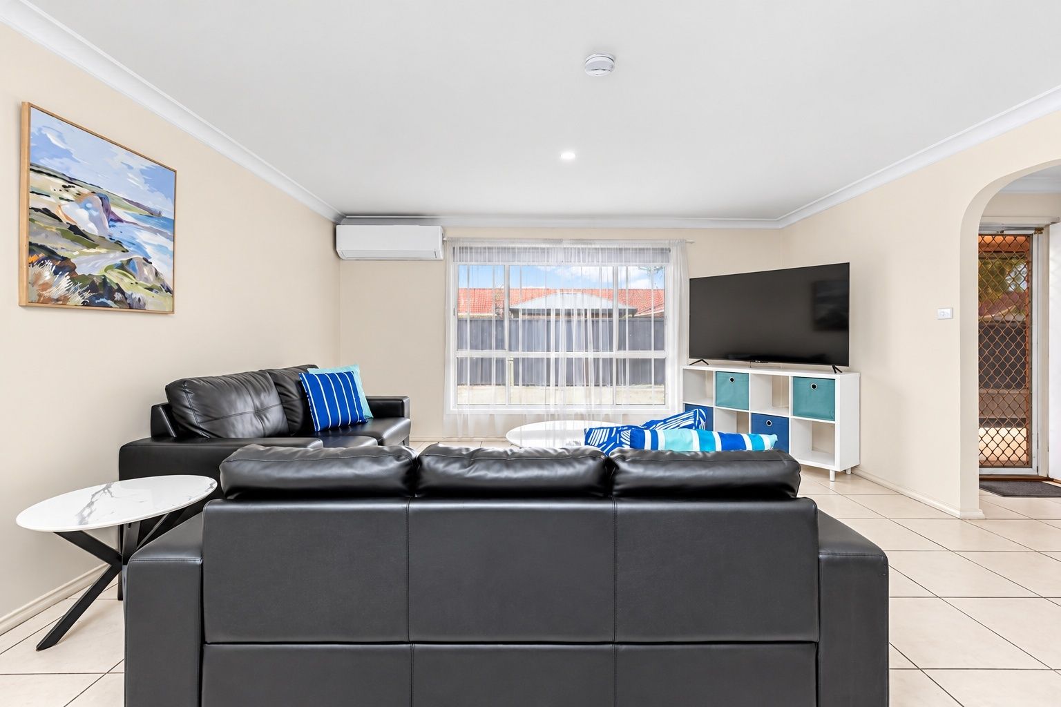 A modern living room with a black leather sofa, a small round table, a flat-screen TV, and a window with blinds.
