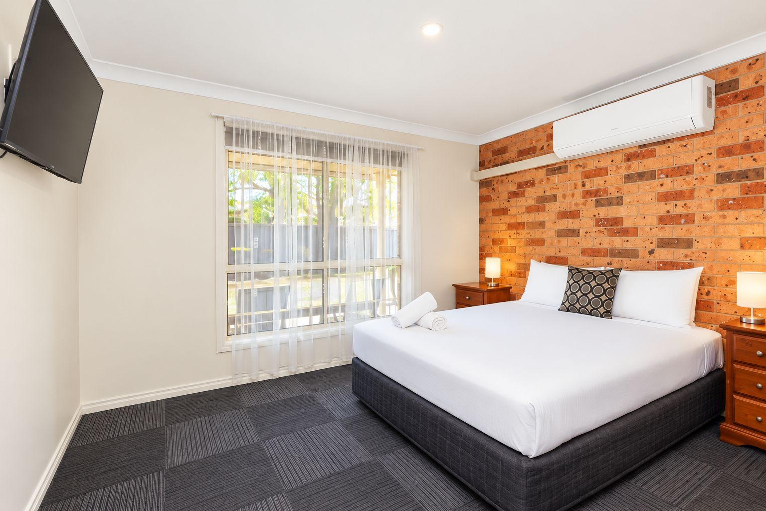 A hotel room with a large bed, a flat-screen TV mounted on the wall, a brick-patterned wall, and a window with sheer curtains.