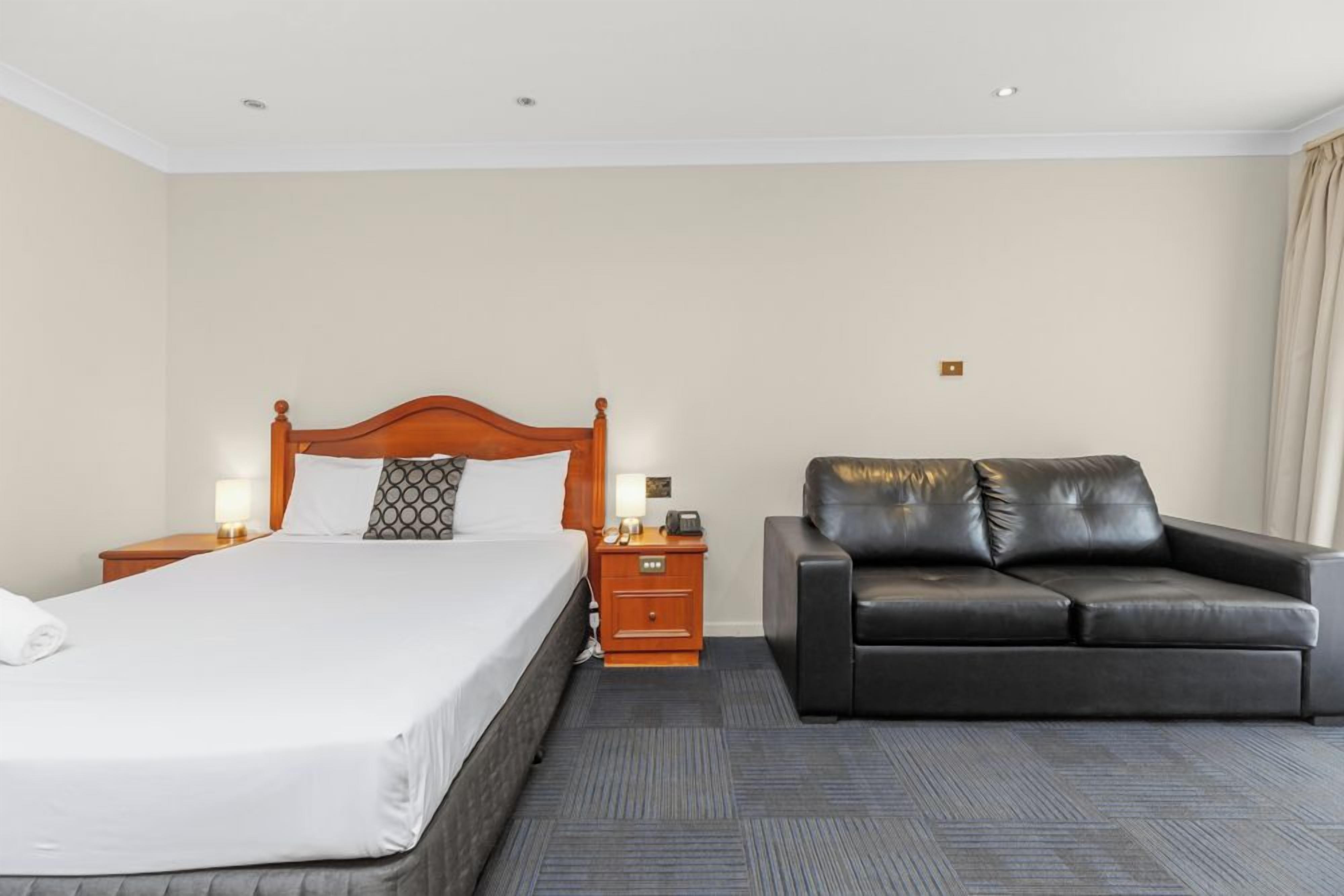 A hotel room with a bed, a nightstand, a lamp, a leather sofa, and a carpeted floor.