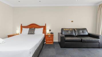 A hotel room with a bed, a nightstand, a lamp, a leather sofa, and a carpeted floor.
