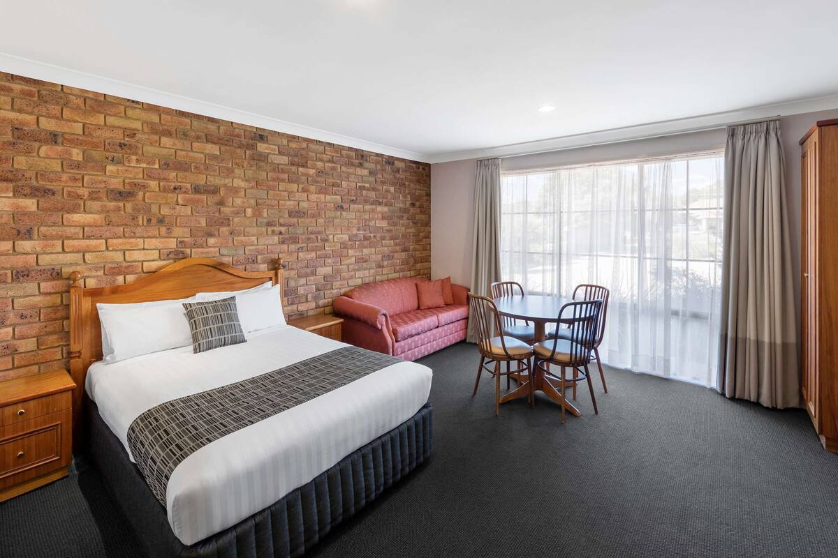 A hotel room with a brick wall, a bed with a wooden headboard, a red sofa, a dining table with chairs, and a large window with curtains.