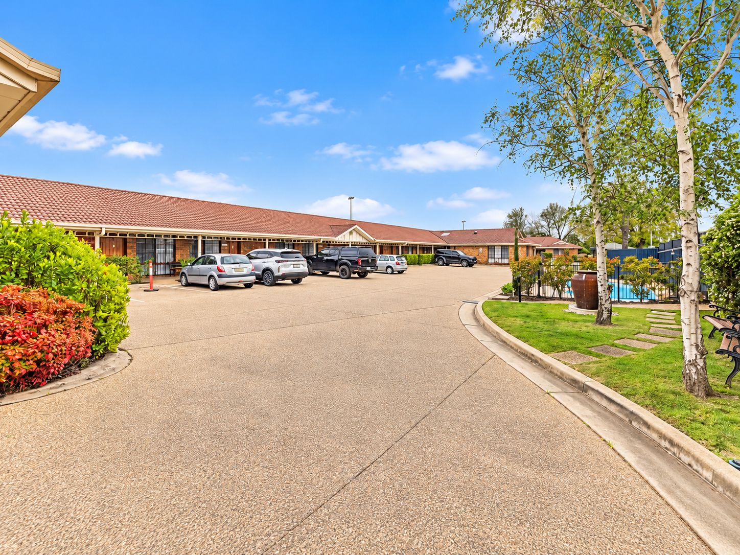 A parking lot with several cars, a building with a red-tiled roof, and a well-maintained lawn with trees and shrubs.