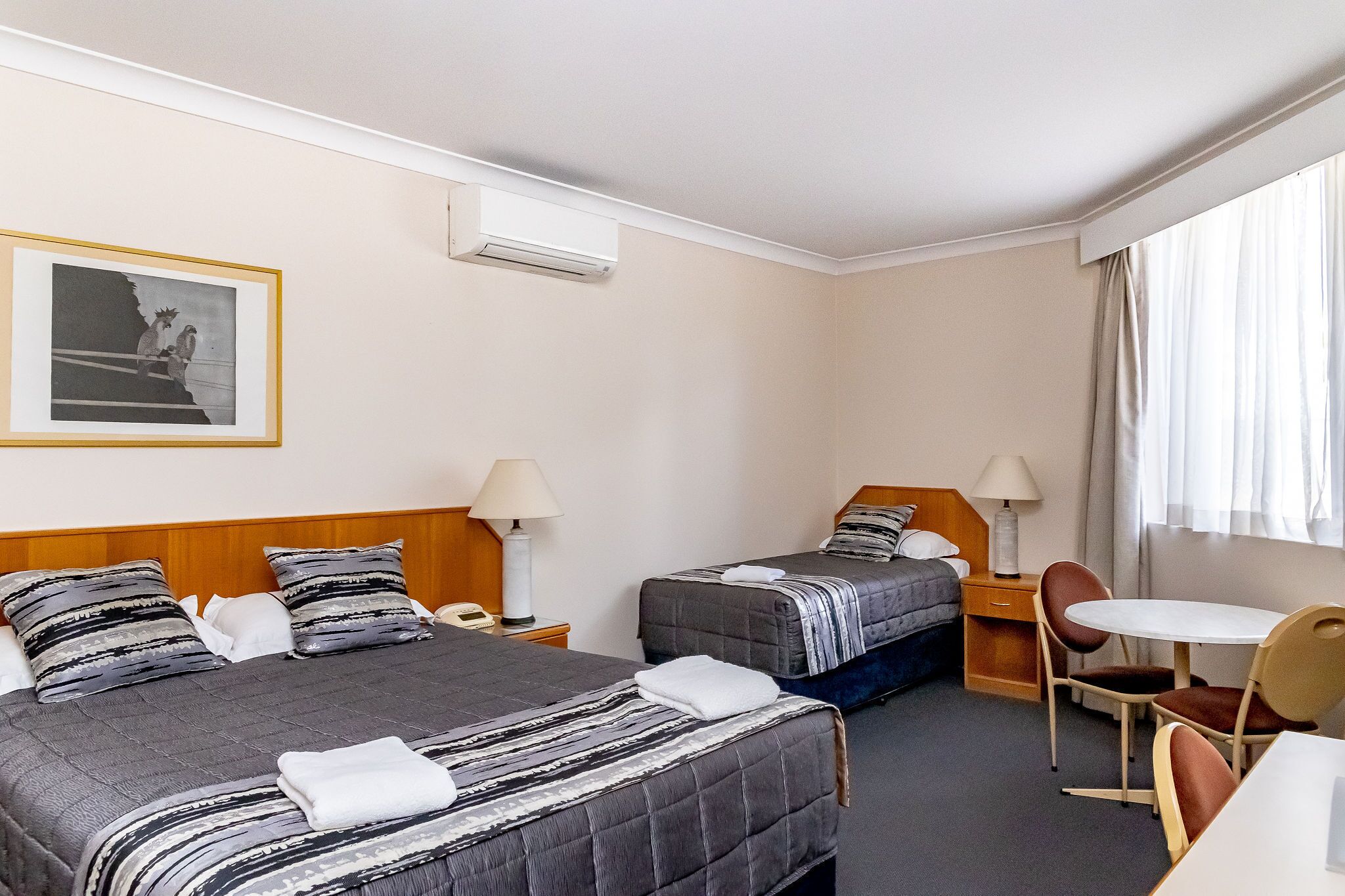 A hotel room with two beds, a small table with chairs, a lamp, and a framed picture on the wall.
