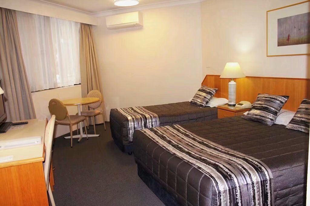 A hotel room with two beds, a desk, a chair, a lamp, and a painting on the wall.