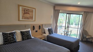 A hotel room with two beds, a small table, a chair, and a balcony with a view of greenery.