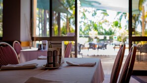 A dining table set with a menu, a bottle, and glasses, with a view of outdoor seating and greenery through large windows. - Charbonnier Motor Inn (Singleton)