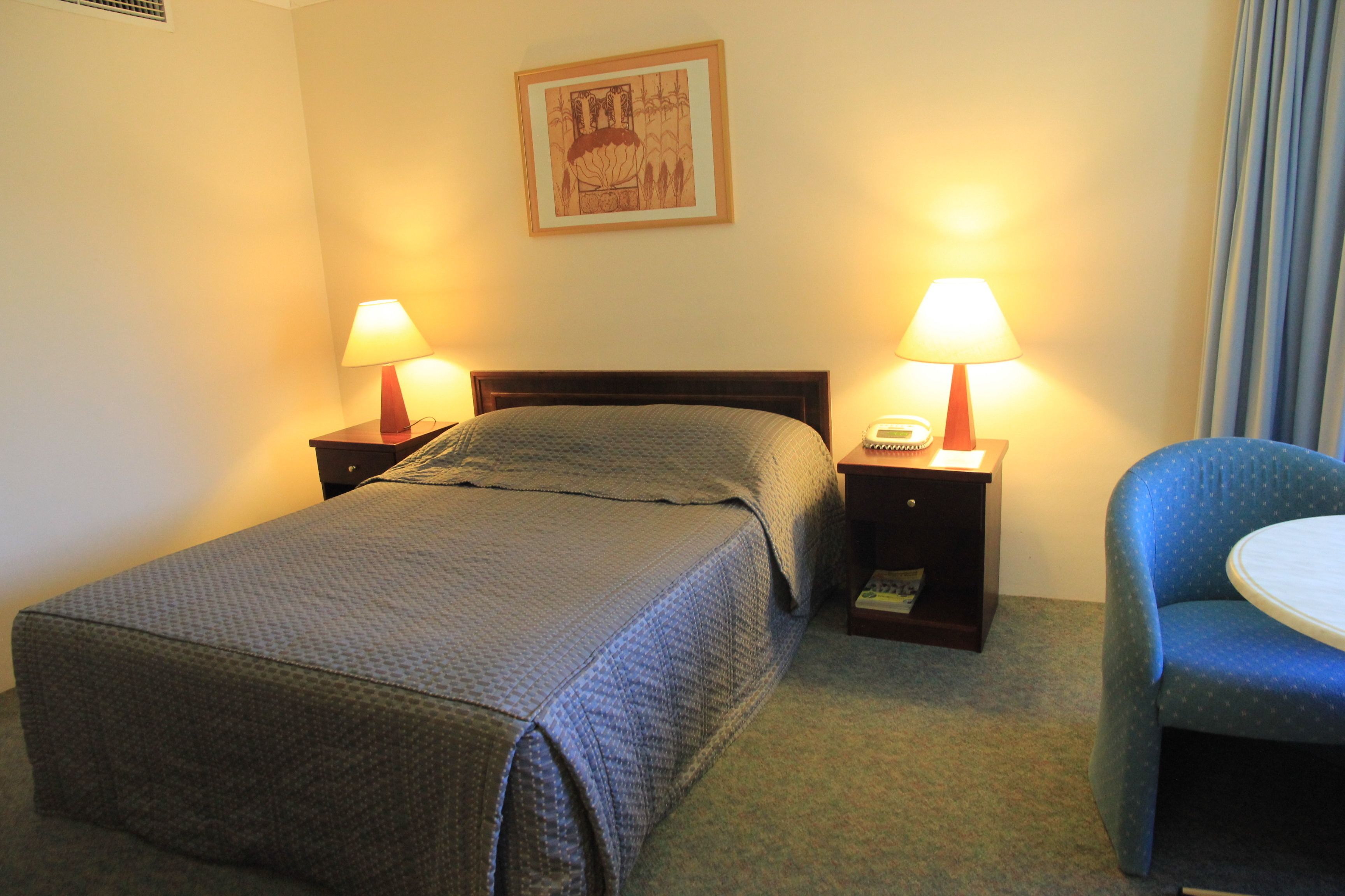 Quarto de hotel com uma cama de solteiro, duas mesas de cabeceira com abajures, uma cadeira e um quadro na parede.