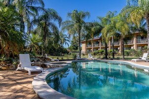 Una piscina con sillones de descanso, rodeada de palmeras y un edificio con balcones.