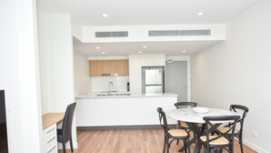 A modern kitchen with white cabinets, a built-in oven, and a refrigerator. There is a dining area with a white table and black chairs. - Brisbane International - Virginia (Boondall)