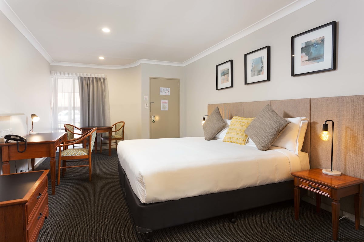 A hotel room with a bed, bedside table, lamp, desk, chair, and a door.