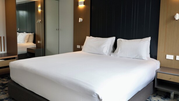 A modern hotel room with a large bed, a bedside table, a nightstand, a mirror, and a wall-mounted light fixture. - Brisbane International - Virginia (Boondall)