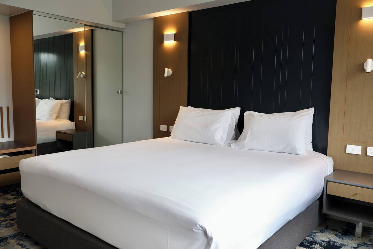 A modern hotel room with a large bed, a bedside table, a nightstand, a mirror, and a wall-mounted light fixture.