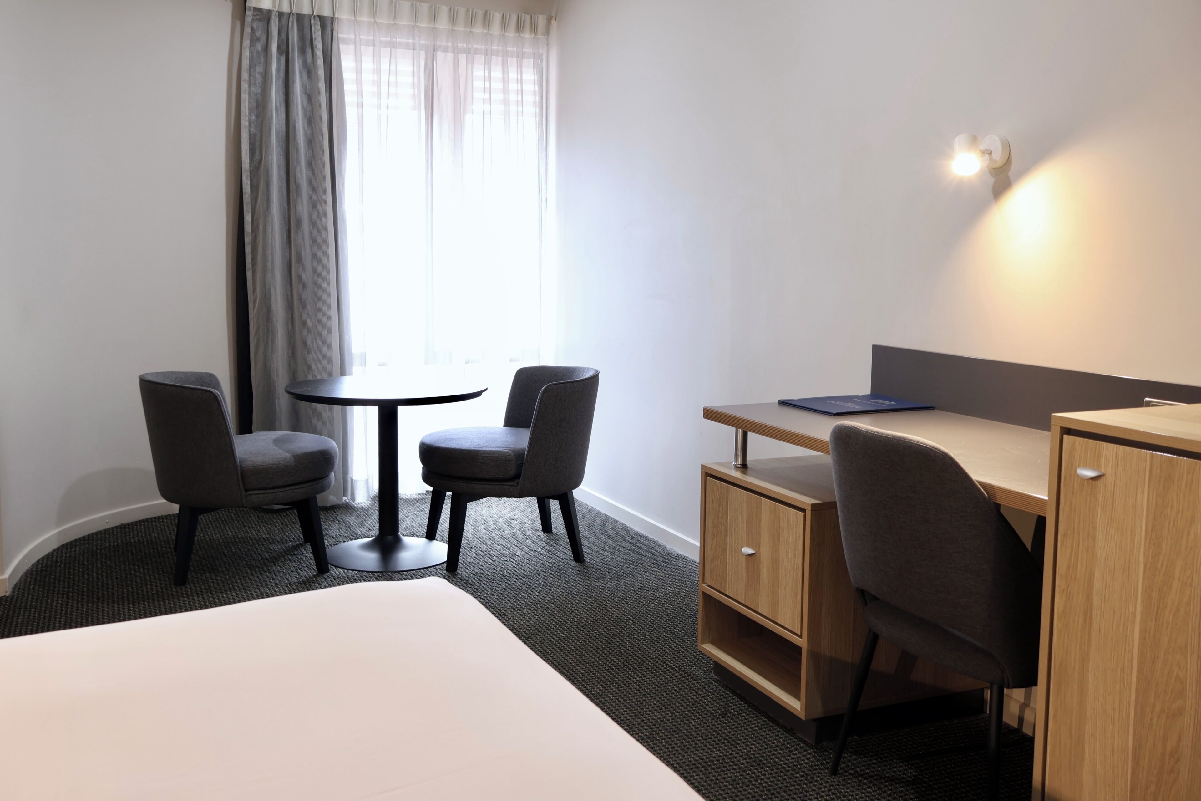 A hotel room with a bed, a desk, two chairs, a small table, and a window with curtains.