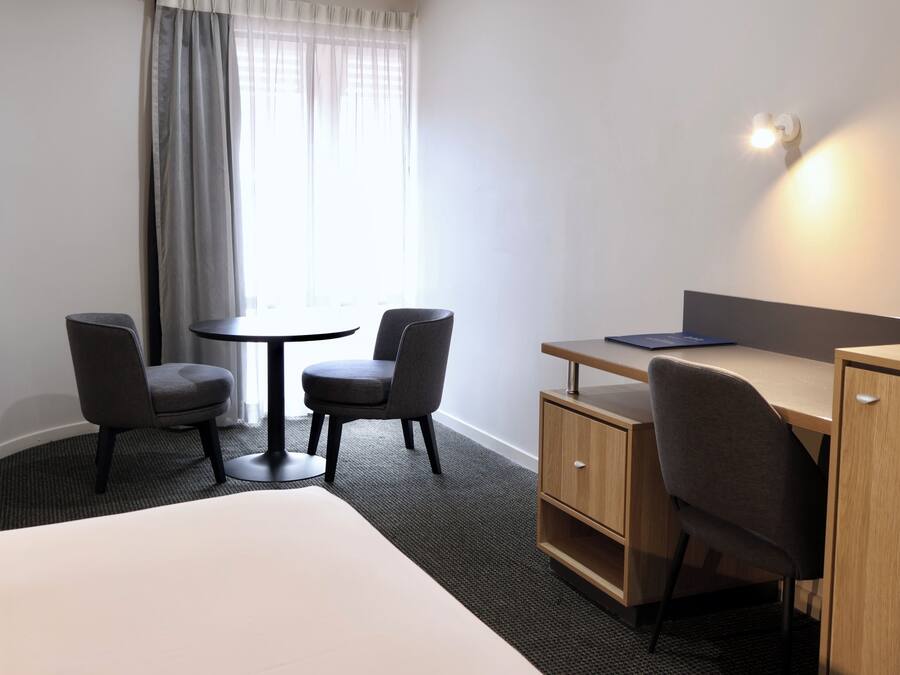 A hotel room with a bed, a desk, two chairs, a small table, and a window with curtains.