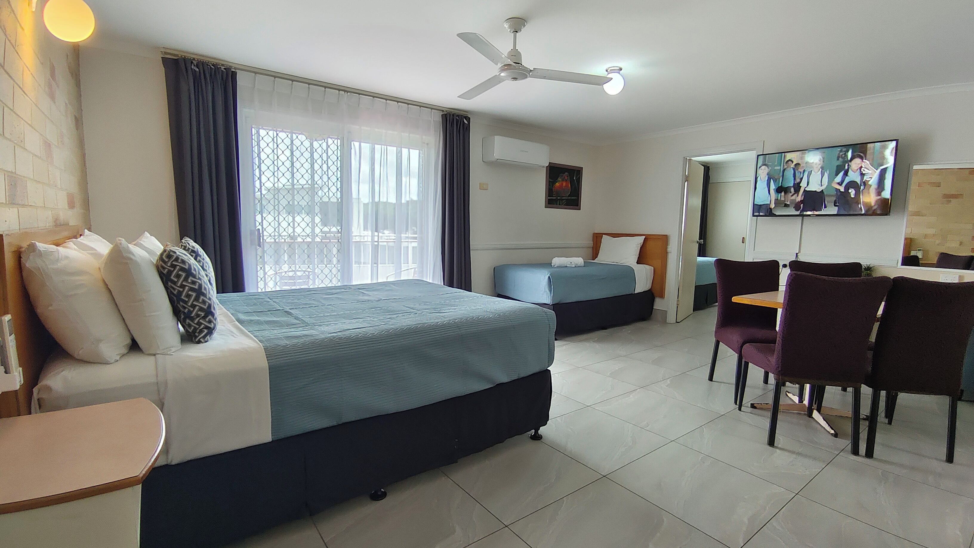 Una camera d'hotel con un letto, una scrivania, una sedia, una TV e una ventola a soffitto.