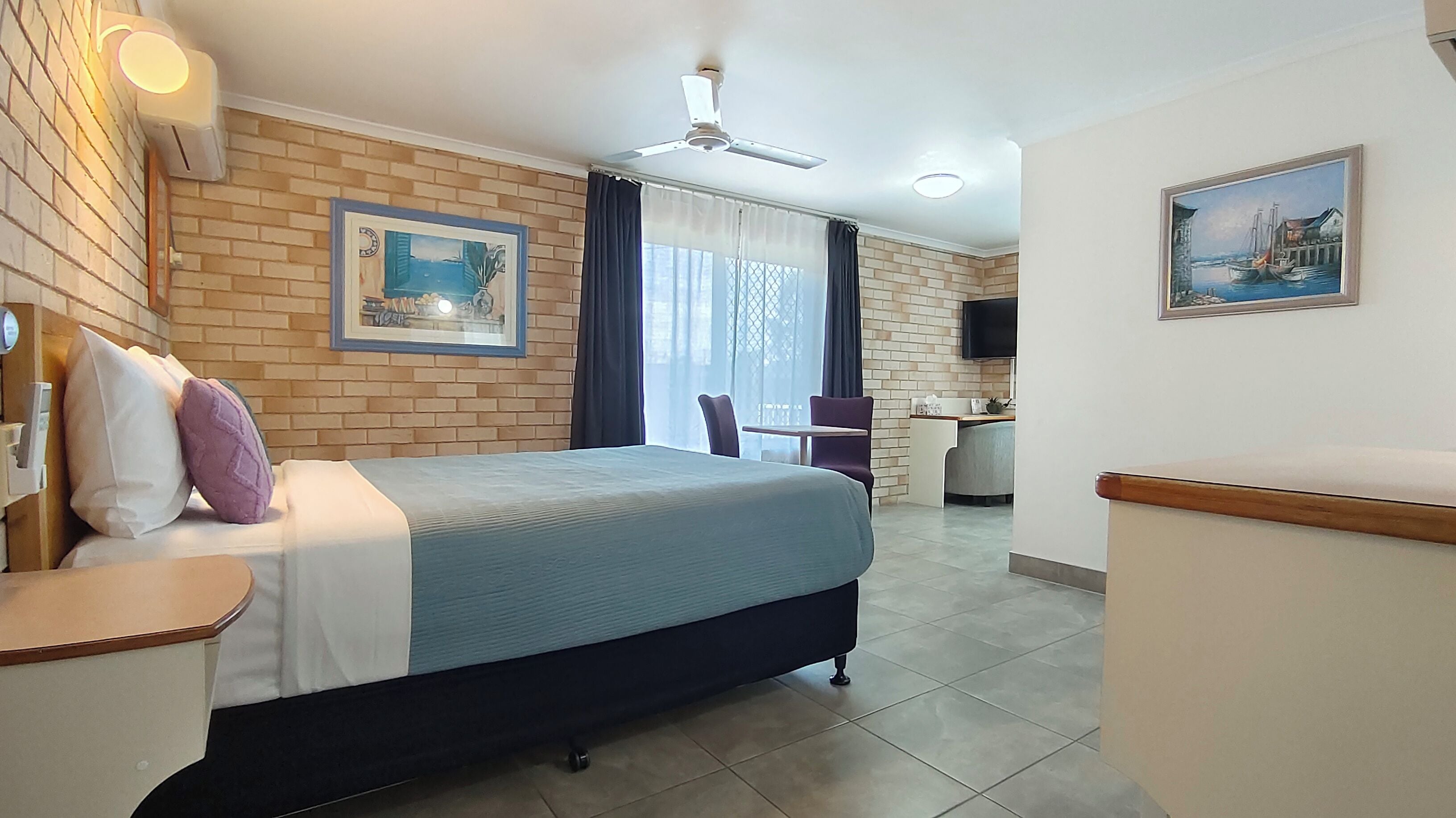 Quarto de hotel com cama, escrivaninha, cadeira, televisão e um quadro na parede.