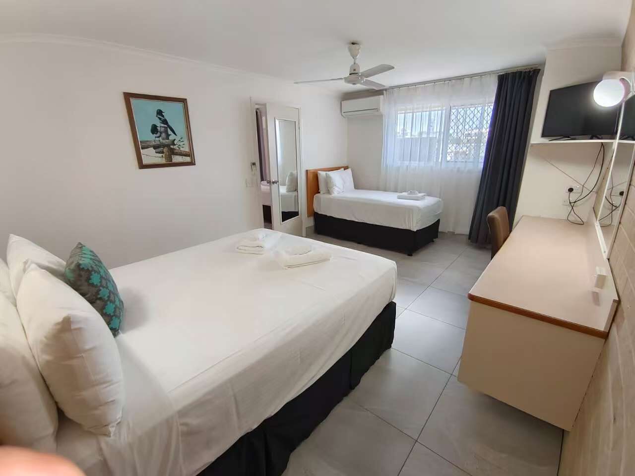 A hotel room with two beds, a desk, a ceiling fan, a TV, and a window with curtains.