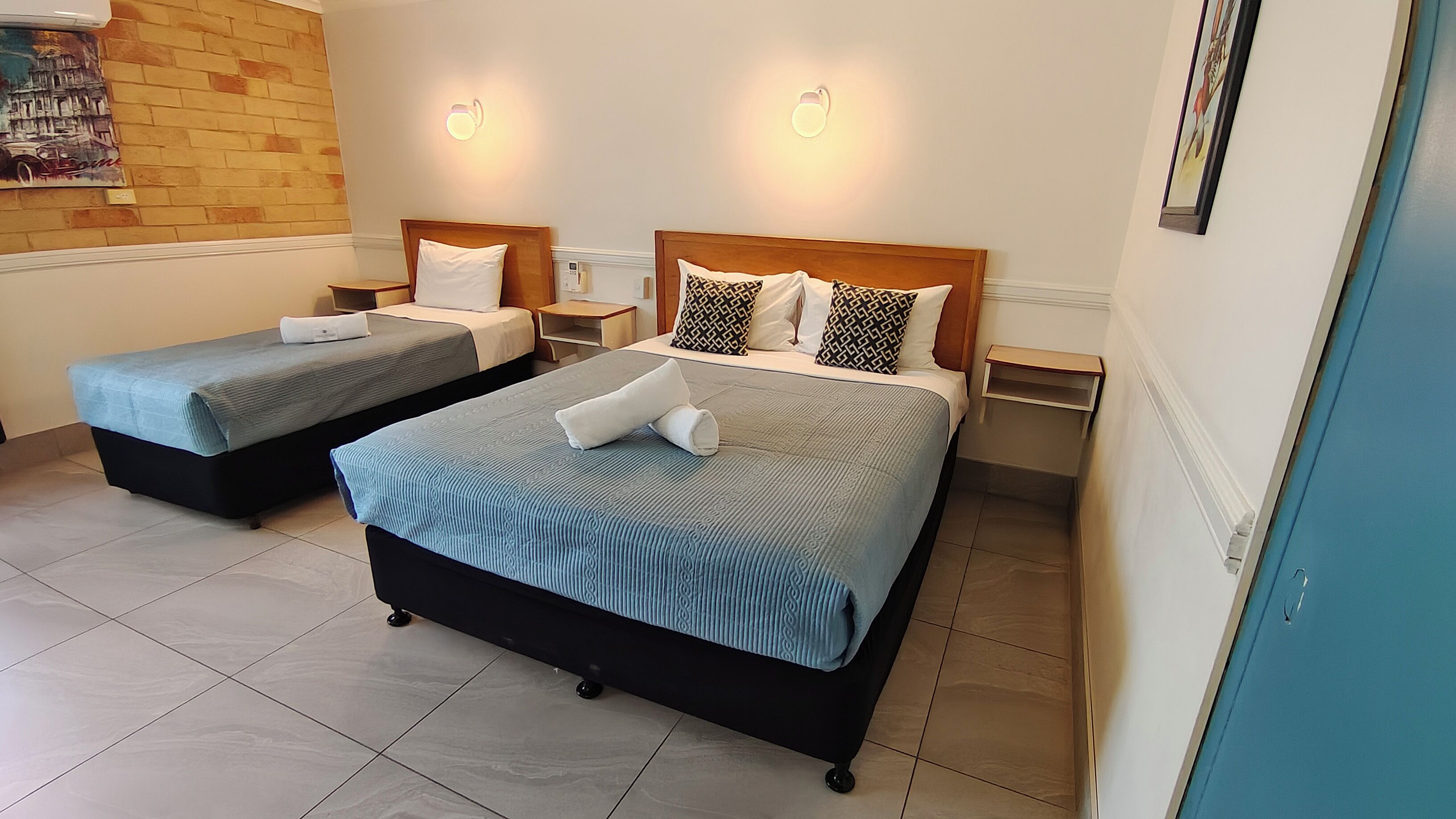 A hotel room with two beds, each with a wooden headboard, bedside tables, and wall-mounted lamps.
