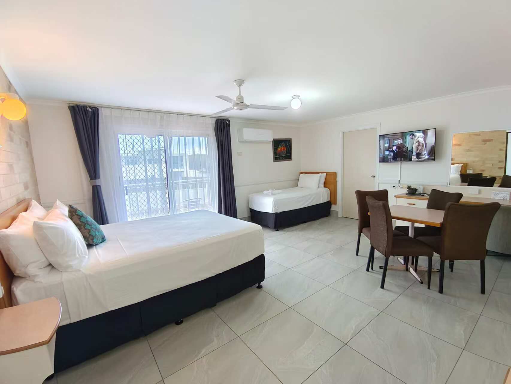 A hotel room with a large bed, a smaller bed, a dining area with chairs, a TV, and a ceiling fan.