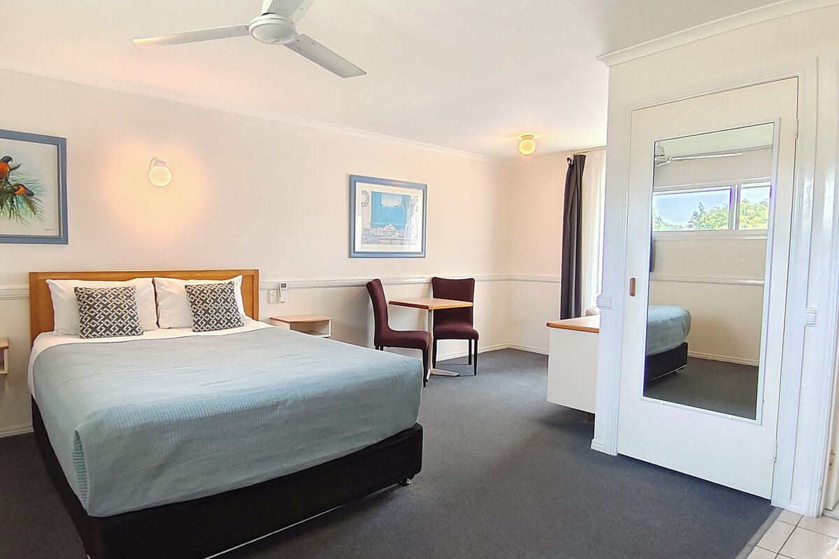 A hotel room with a bed, a desk, a chair, a ceiling fan, and artwork on the walls.