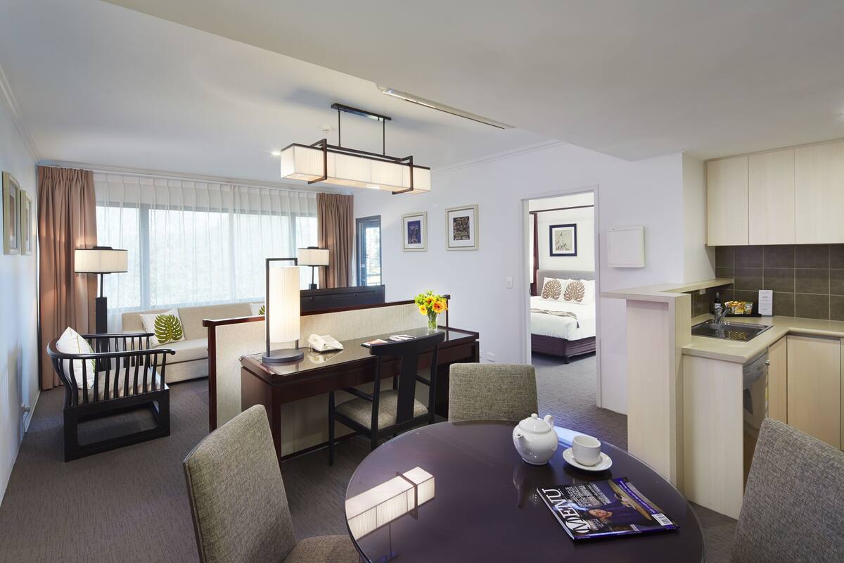 A modern hotel room with a dining area, a kitchenette, and a separate bedroom visible through an open door.