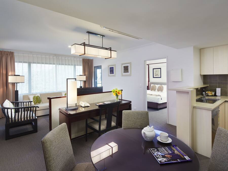 Une chambre d’hôtel moderne dotée d’un coin repas, d’une kitchenette et d’une chambre à coucher séparée, visible à travers une porte ouverte.