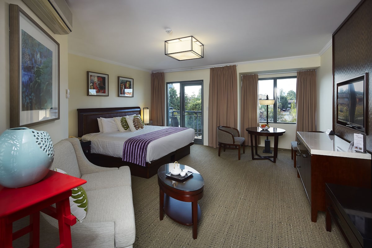 A hotel room with a bed, a desk, a chair, a TV, and a balcony with curtains.