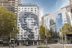 A hotel with a large mural on its facade, a modern glass building adjacent to it, and a street intersection with traffic lights.