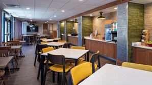 Un restaurante con mesas de madera y sillas amarillas, una barra con dispensador de bebidas y una pantalla de televisión.