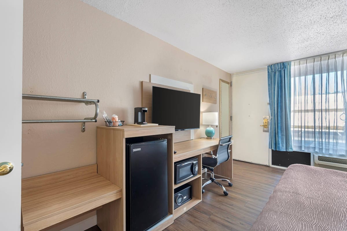 A hotel room with a wooden desk, a flat-screen TV, a mini-fridge, a microwave, a chair, and a bed.