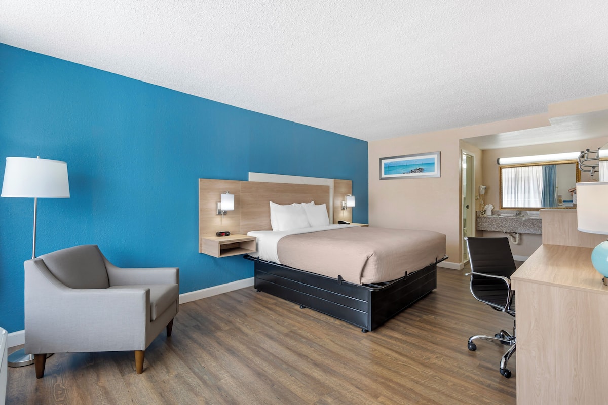 A hotel room with a bed, a desk, a chair, a lamp, and a nightstand.