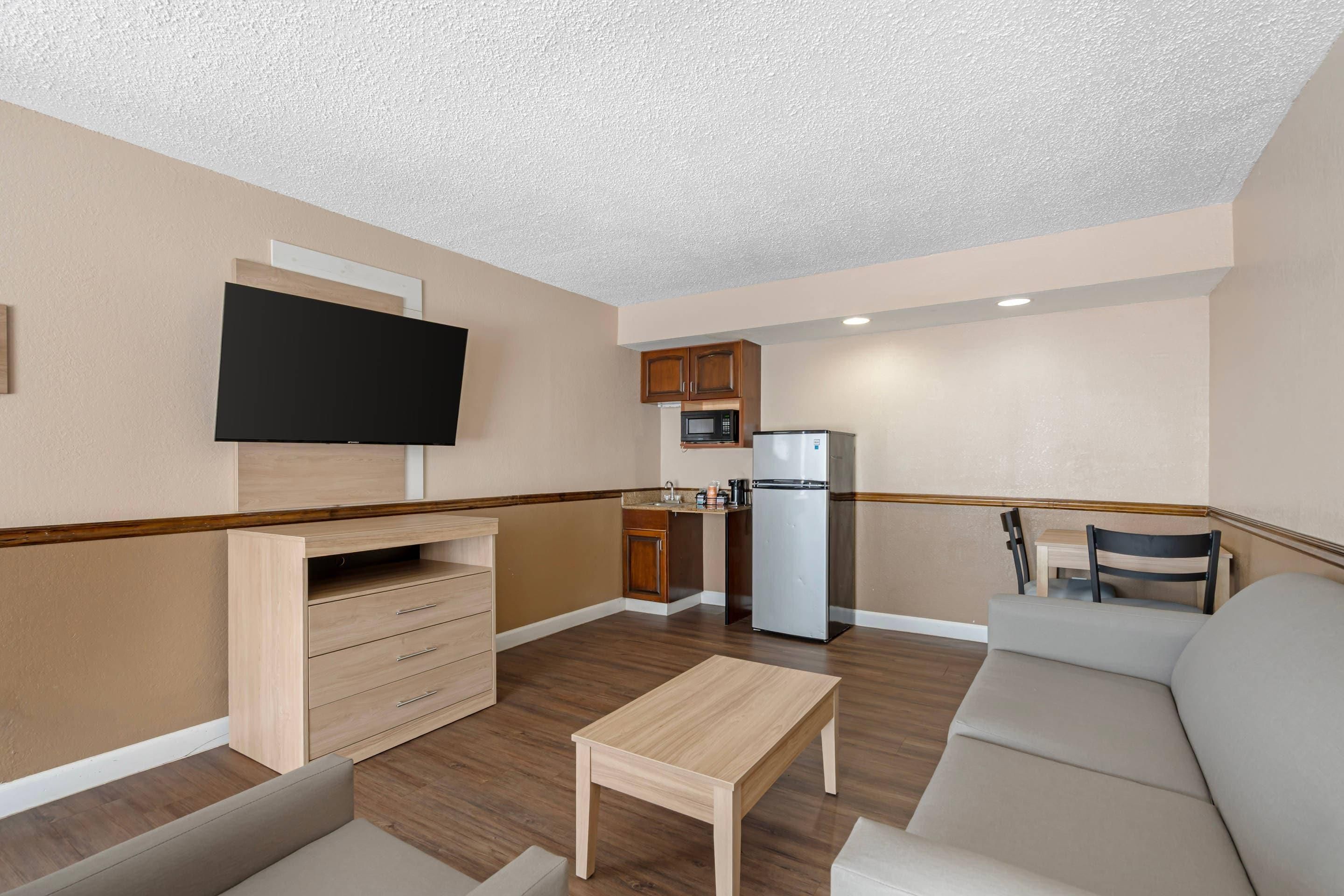 A compact living space with a kitchenette, a flat-screen TV mounted on the wall, a sofa, a coffee table, and a small dining table.