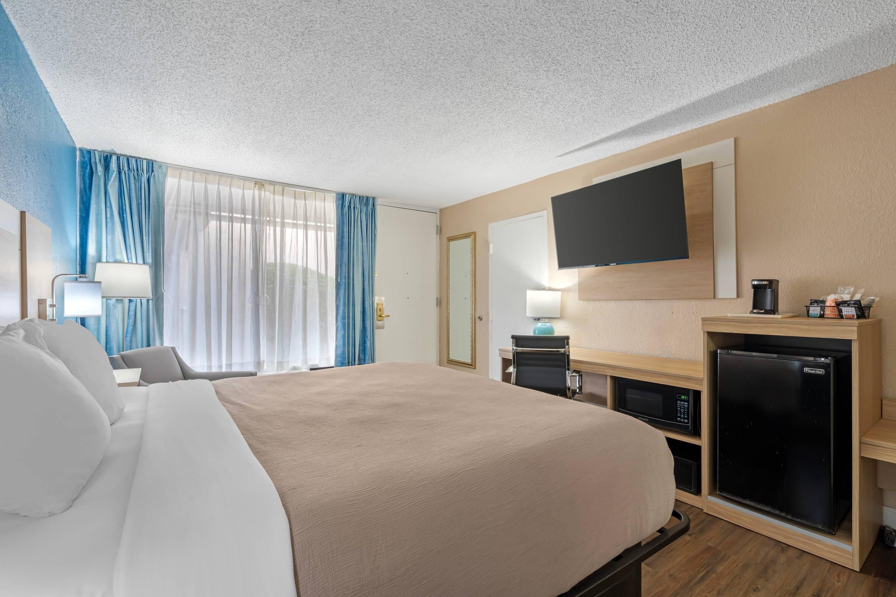 Ein Hotelzimmer mit einem großen Bett, einem flachbildfernseher an der Wand, einem Schreibtisch mit Computer und Kaffeemaschine sowie einem Minikühlschrank.