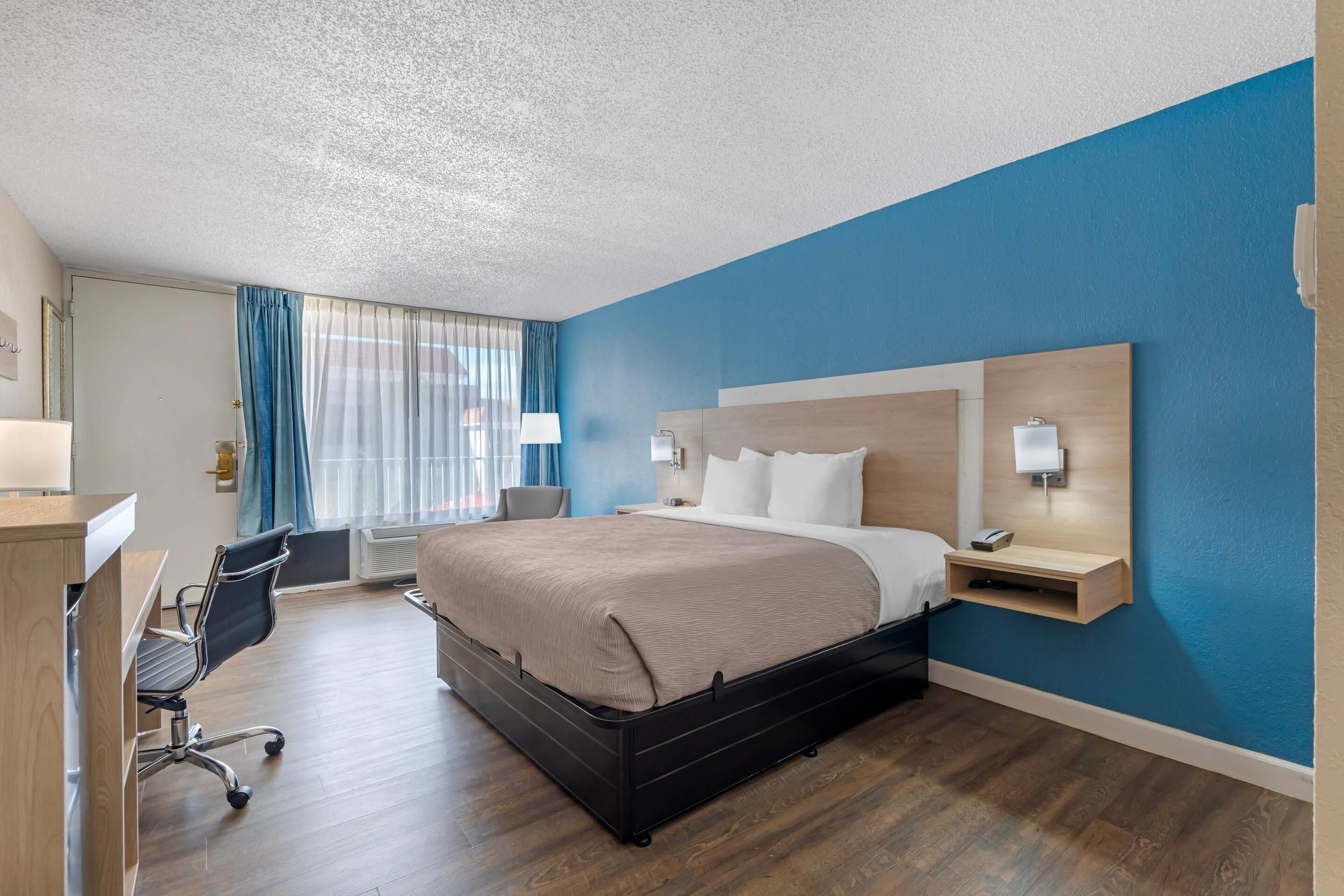 A hotel room with a large bed, a desk, a chair, and a window with curtains.