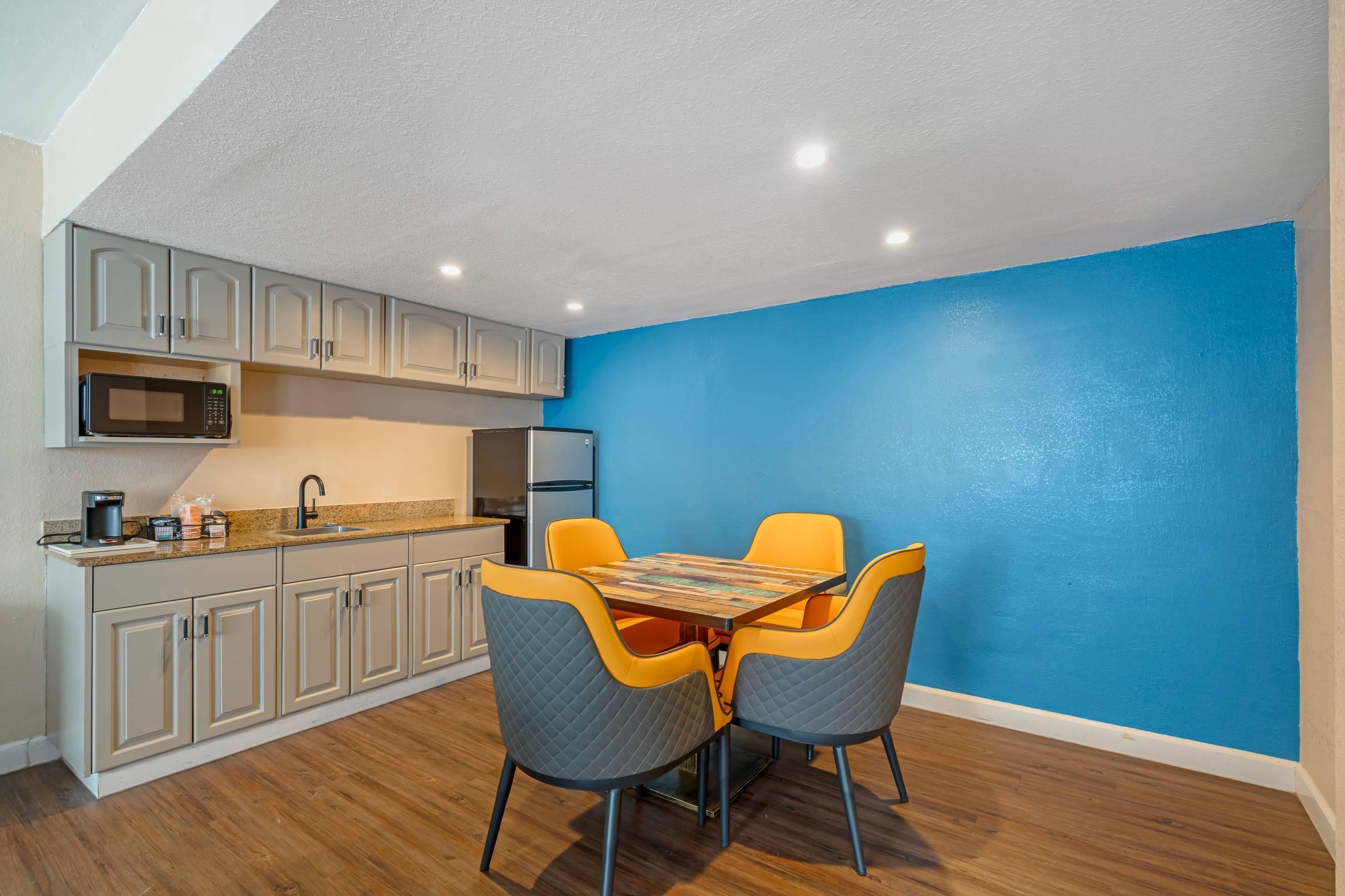 A compact kitchen with a microwave, refrigerator, and a coffee maker. There is a dining table with four chairs, and the walls are painted blue.