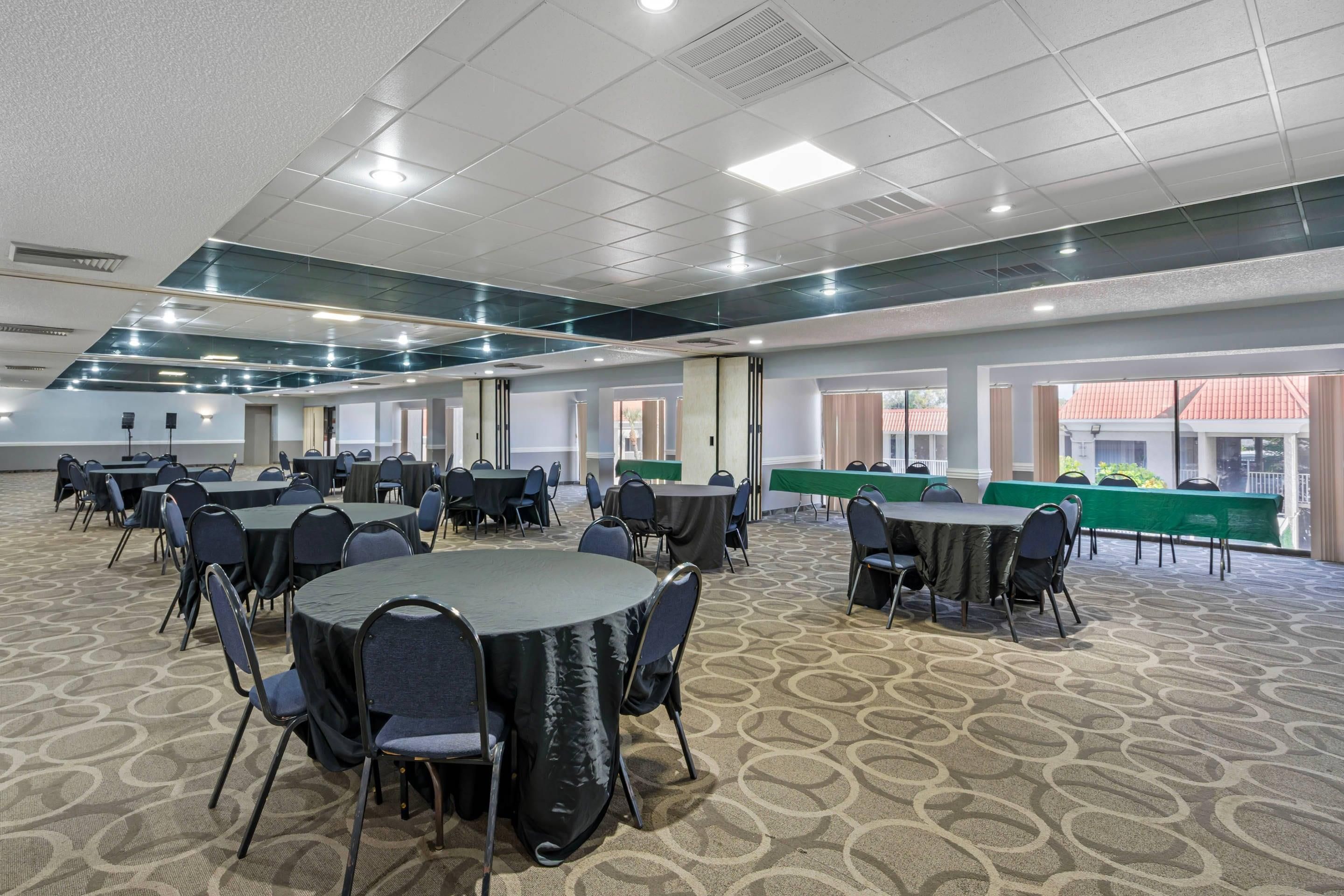 A large conference room with multiple round tables and chairs, carpeted floor, and large windows with views of buildings outside.