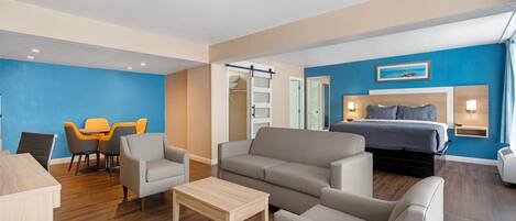 A modern hotel room with a sofa, armchairs, a small table, a bed, and a desk area with chairs.