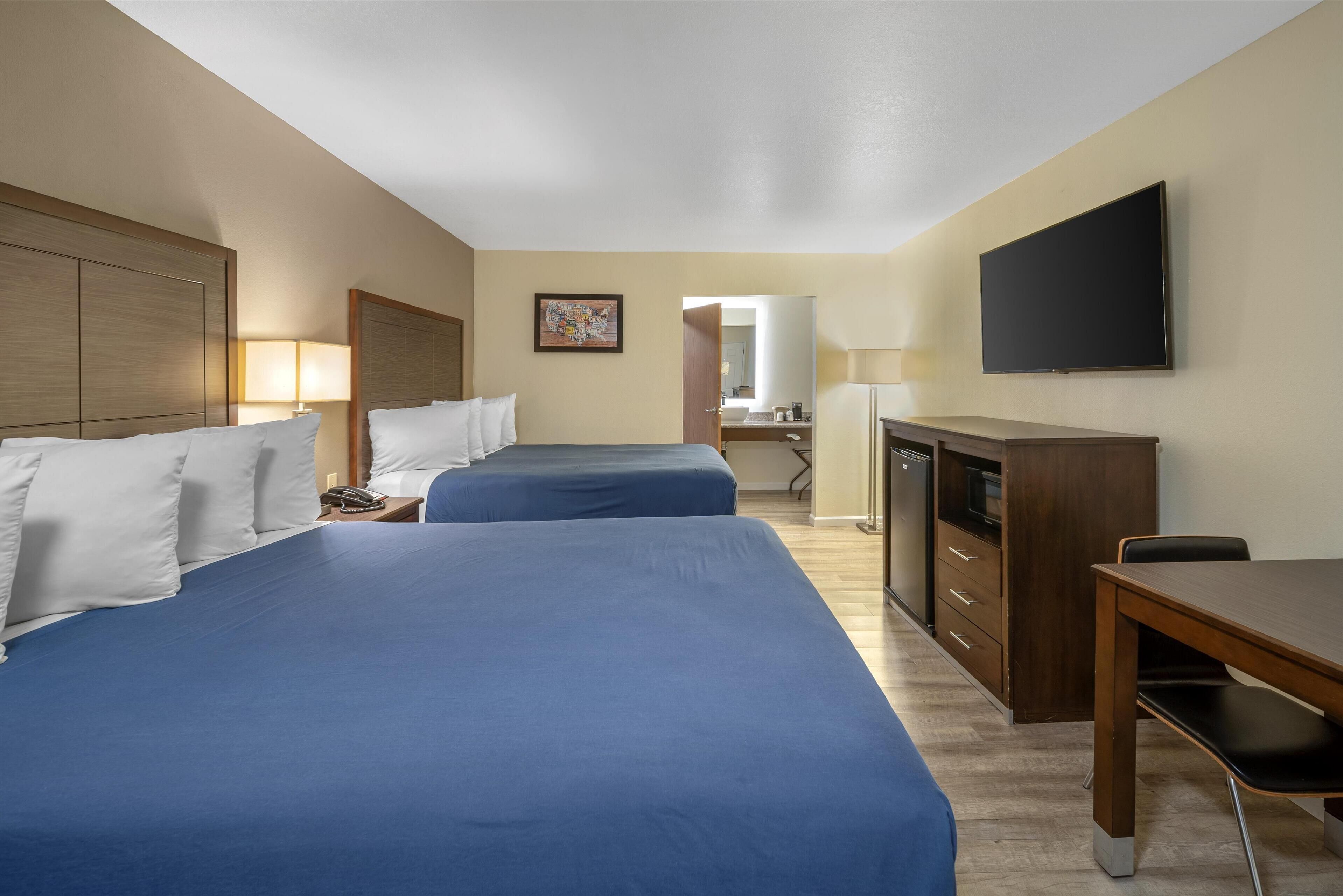 A hotel room with two beds, a desk, a TV, a mirror, and a bathroom visible in the background.