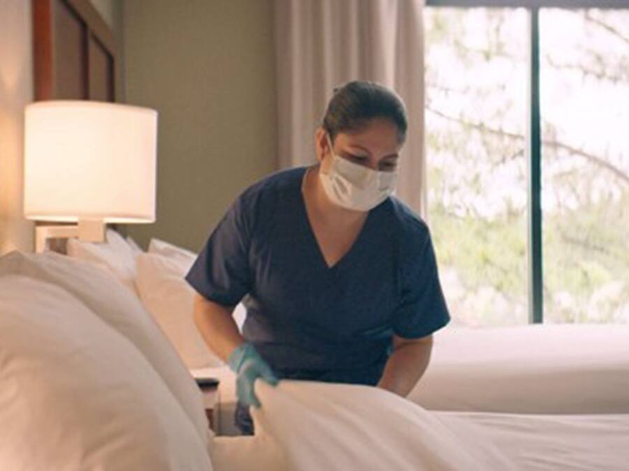 A person in a blue uniform cleaning a bed in a room with a window and a lamp.