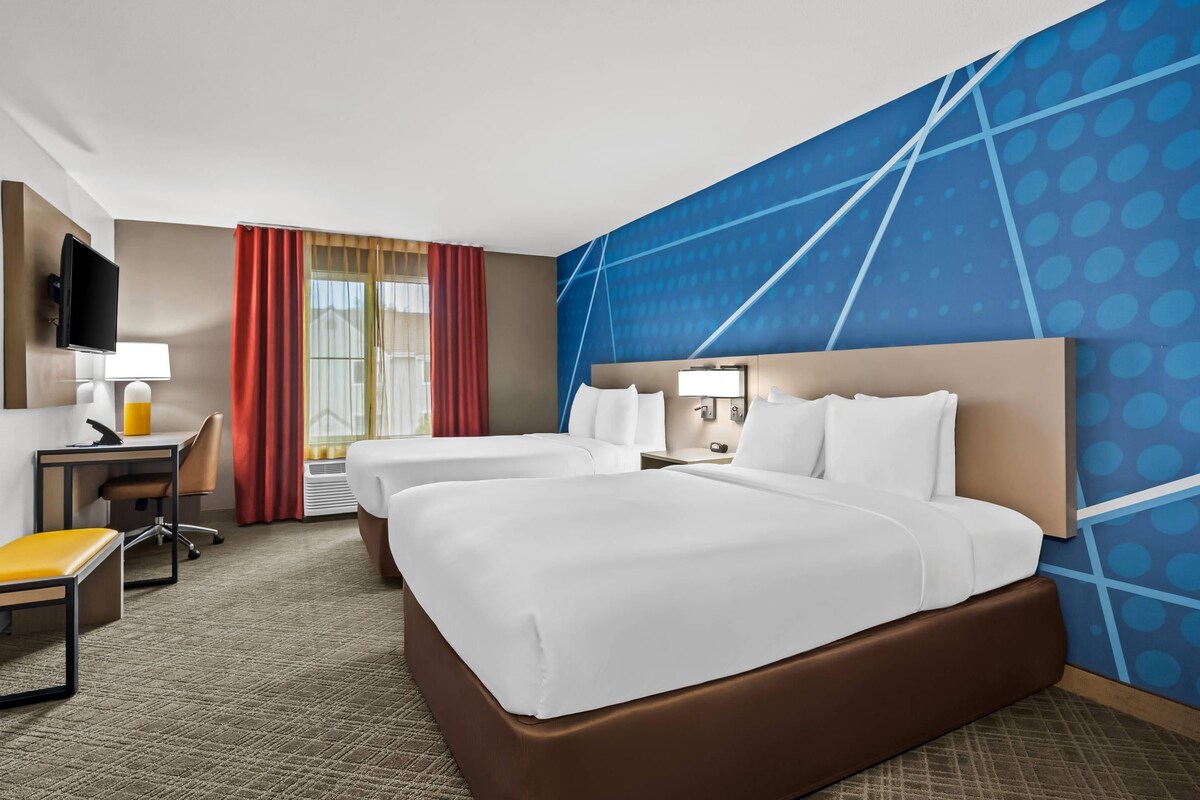 A hotel room with a large bed, two bedside tables, a desk with a chair, a TV, and a window with curtains.